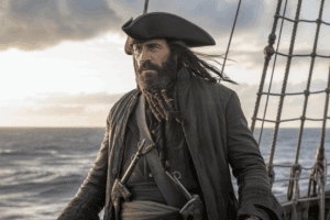 Ein schroffer Pirat mit langen dunklen Haaren, Dreispitz und Pistolen steht auf dem Deck eines Schiffes auf See, mit dem Meer und einem wolkenverhangenen Sonnenuntergangshimmel im Hintergrund.