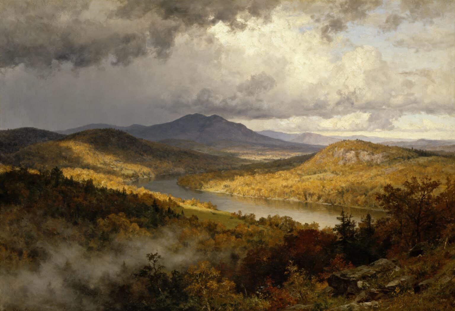 Ein Fluss schlängelt sich durch eine sonnenbeschienene Herbstlandschaft mit bunten Bäumen, sanften Hügeln und fernen Bergen unter einem dramatisch bewölkten Himmel. Aus dem bewaldeten Tal im Vordergrund steigt Nebel auf.