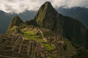 Die alten Steinruinen von Machu Picchu liegen auf einem grünen Bergrücken, umgeben von üppigen, nebligen Gipfeln und einem wolkenverhangenen Himmel, der die atemberaubende Schönheit Perus zeigt.